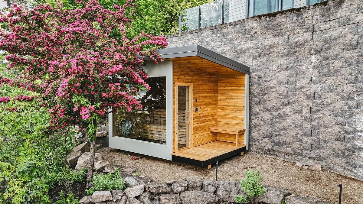 Finished 8x12 Mode Sauna with cedar interior and full glass front beside a flowering tree, Okanagan Lake, BC