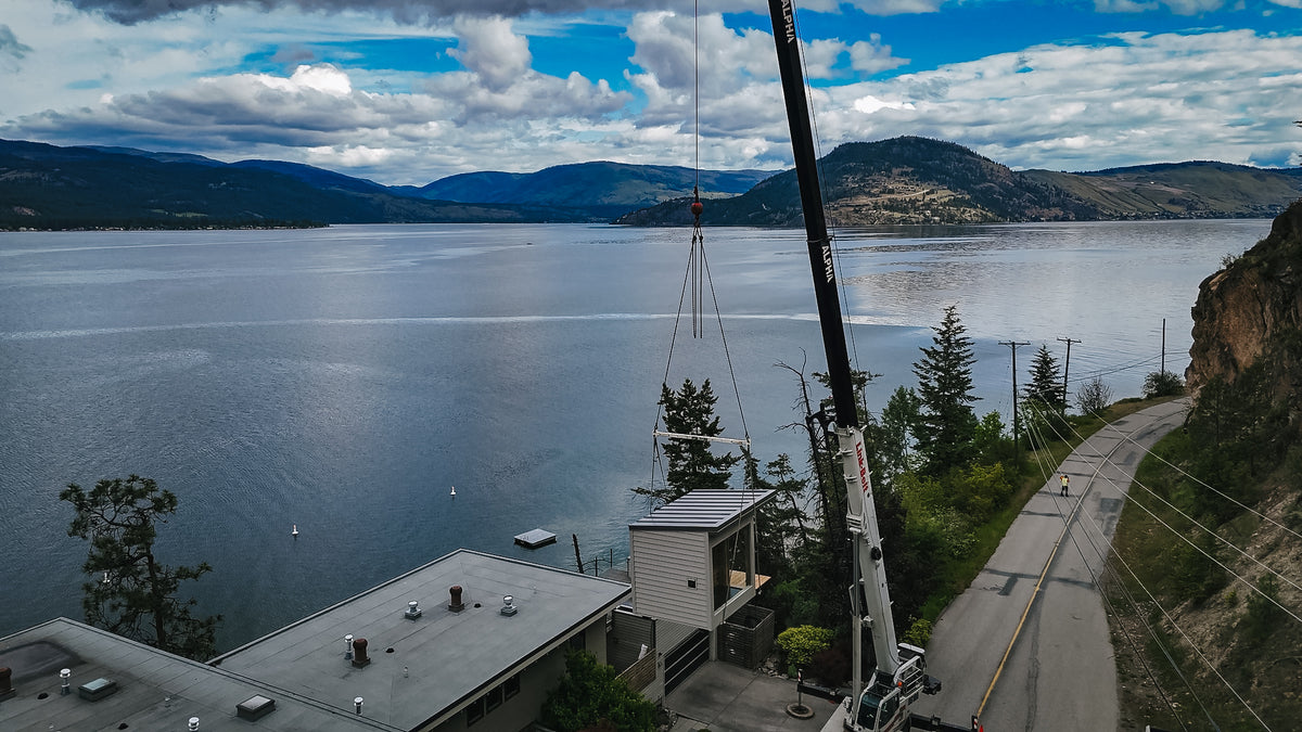Alpha Cranes lifting a finished modular sauna over a house with Okanagan Lake in the background