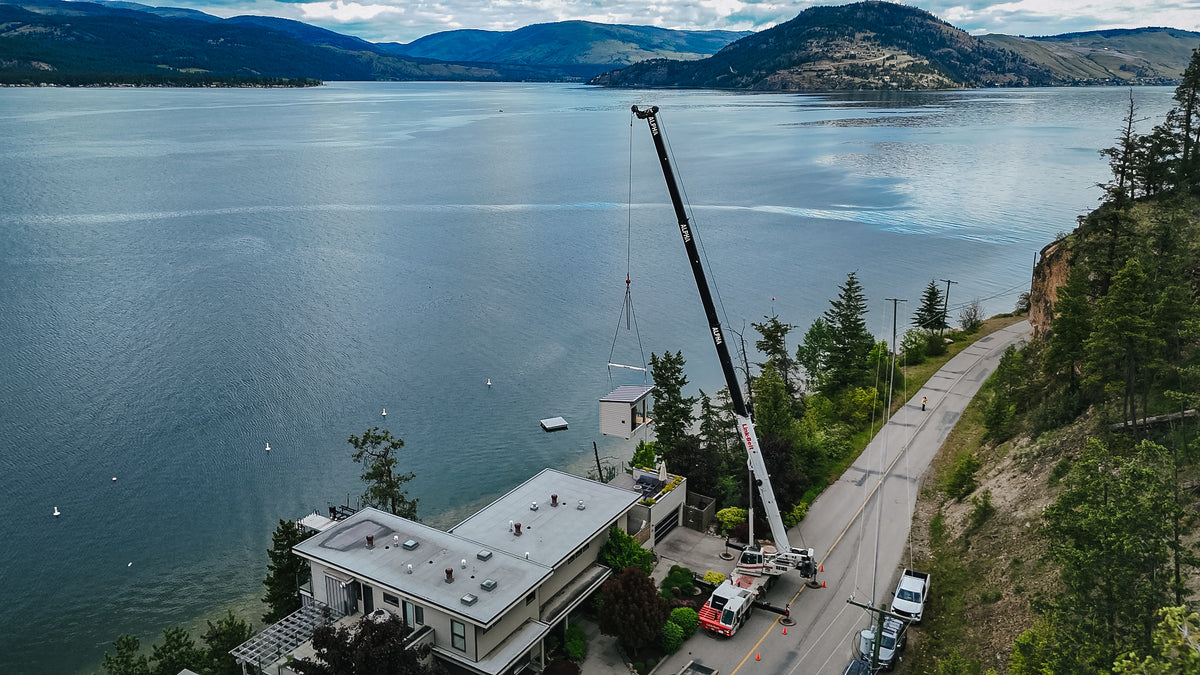 Crane boom extended over a lakefront home during a modular sauna delivery