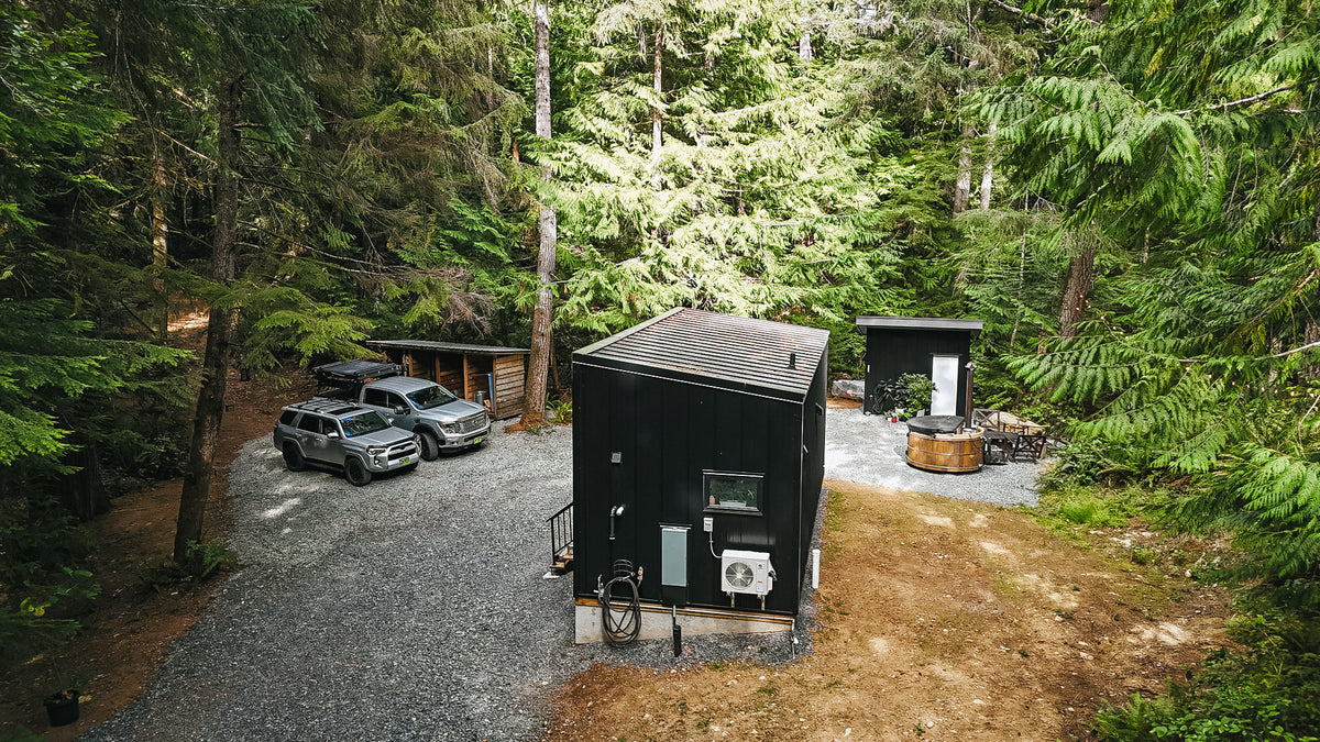 Drone view of the rear and side of a black modular cabin on a gravel pad in the forest